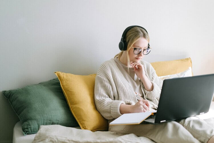 Young blonde girl with glasses sitting at her laptop writing in a notebook