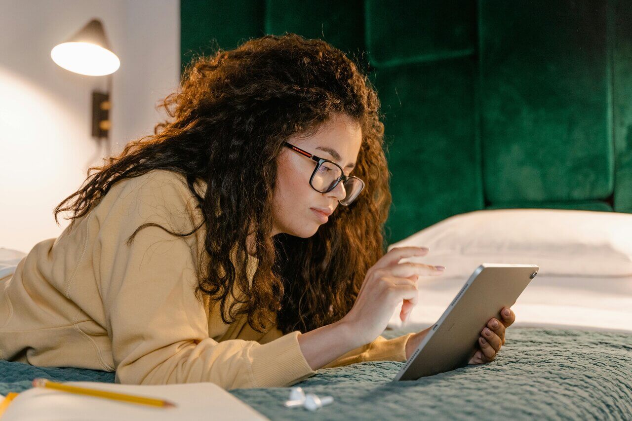 A young woman looking for job interviews on her tablet while lying in bed.